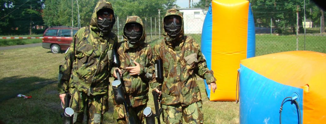 Trzy osoby ubrane w stroje paintballowe stoją przy dmuchanych ściankach na imprezie plenerowej.