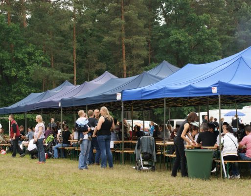 Ludzie siedzą pod namiotem, przy stołach na pikniku zorganizowanym na polanie.