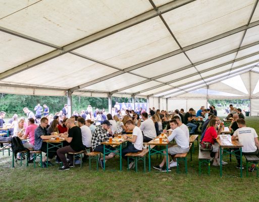 Ludzie siedzą przy stołach pod namiotem na firmowej imprezie w plenerze.