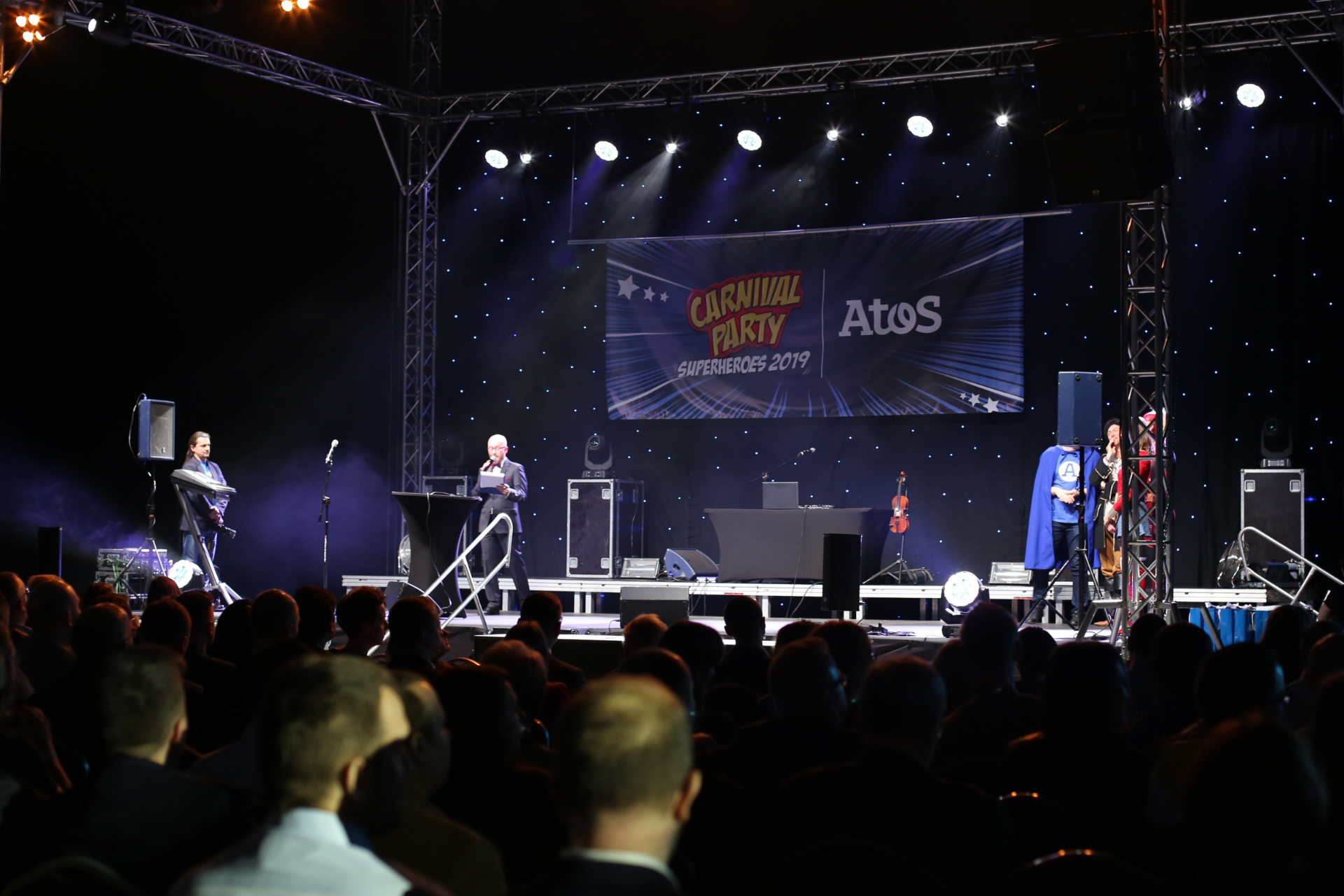 Oświetlona scena z konferansjerem i ludźmi w trakcie imprezy integracyjnej.